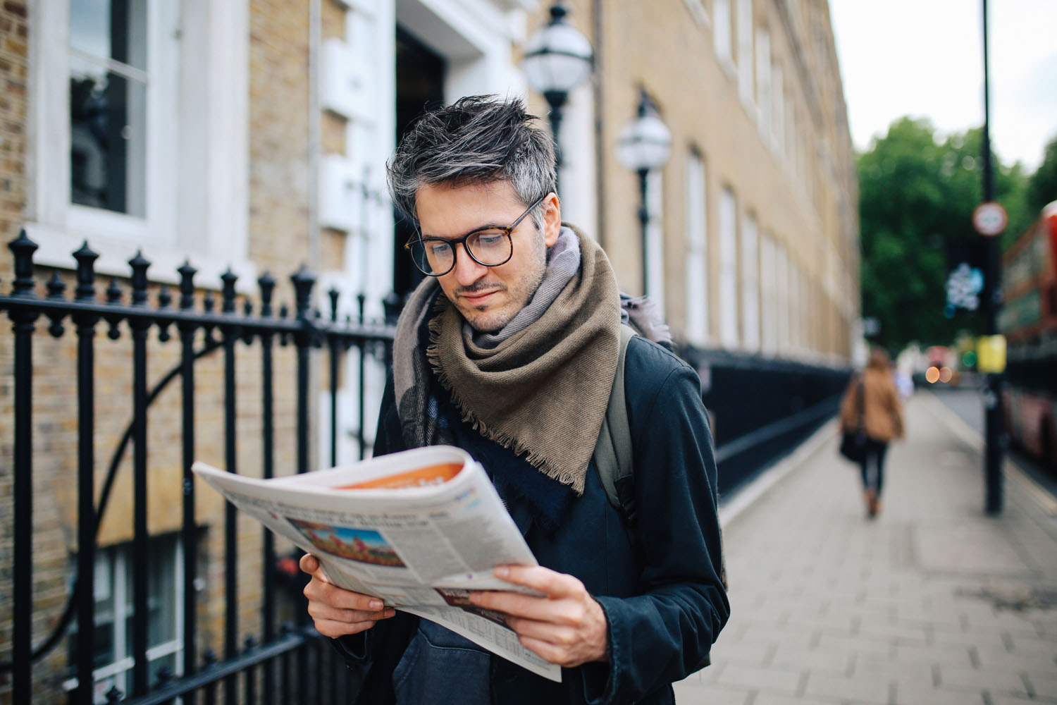 Mann mit Schal ließt eine Zeitung während er auf einer Straße geht.